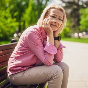 Westside Behavioral Care Depression, Anxiety Section - Thoughtful older woman in glasses and a pink shirt sitting on a park bench, resting her face in her hands with a gentle expression.