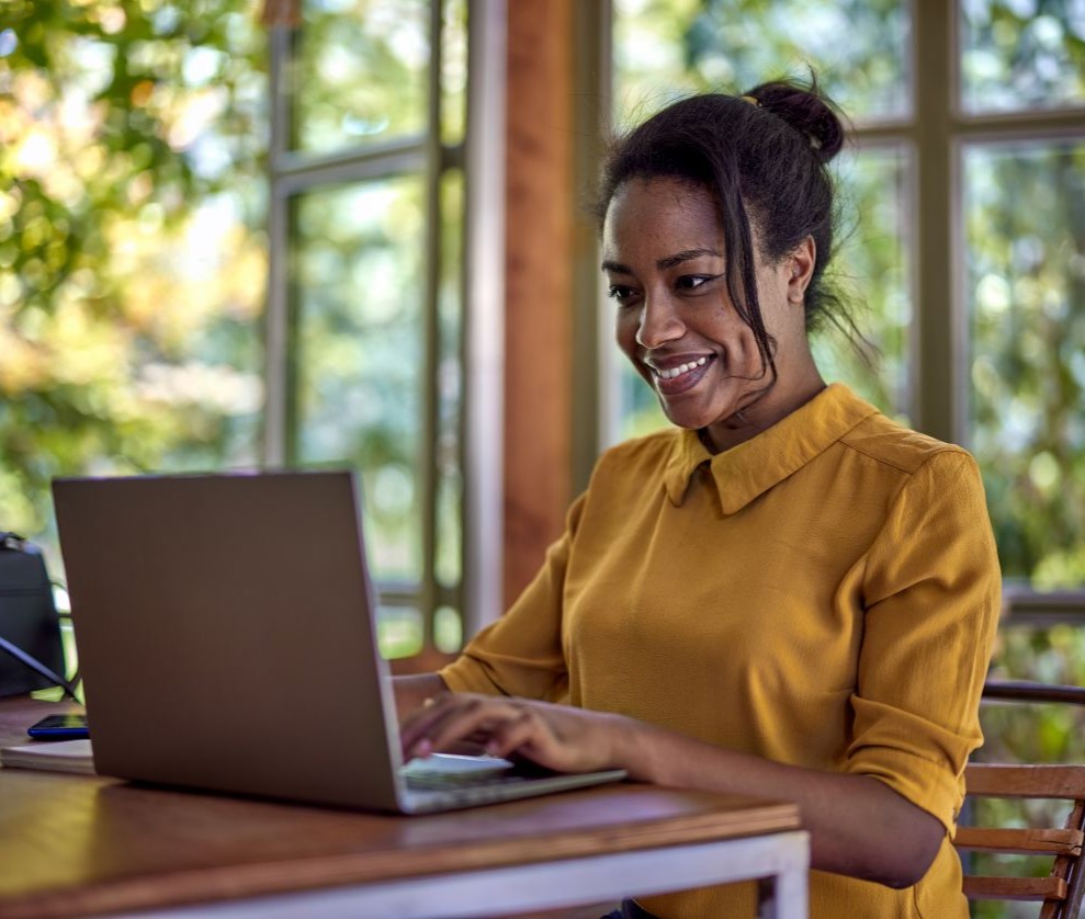 Westside Behavioral Care We Accept Insurance Section - Woman wearing a mustard yellow shirt smiling while working on a laptop at a wooden table in a bright room with large windows and green trees outside.